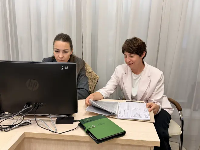 Иллюстрация из медиатеки ГРЦ. Сотрудники ГРЦ - девушка в сером костюме сидит за столом и работает за компьютером, вторая девушка в белом пиджаке сидит рядом и рассматривает документы.